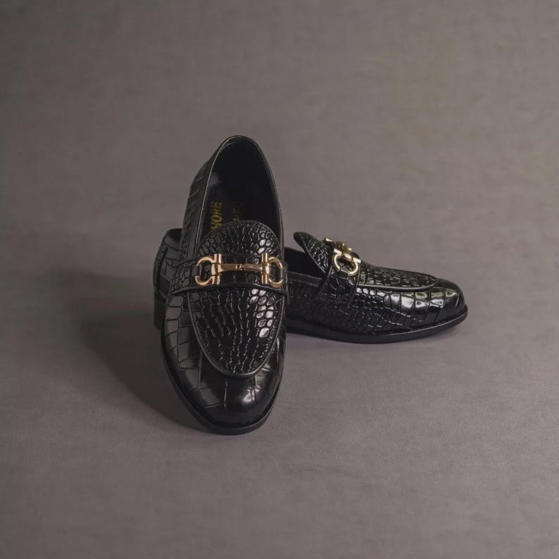K&ouml;nnte beinhalten: Ein Paar schwarze Leder-Loafer mit Krokodiltextur. Die Schuhe haben eine goldfarbene Metallschnalle am Blatt. Die Schuhe werden auf grauem Hintergrund pr&auml;sentiert.