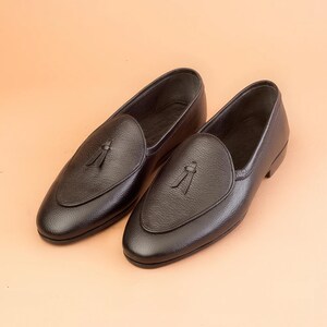 May include: A pair of black leather loafers with a tassel detail on the vamp. The shoes have a rounded toe and a low heel. The loafers are displayed on a peach-coloured background.