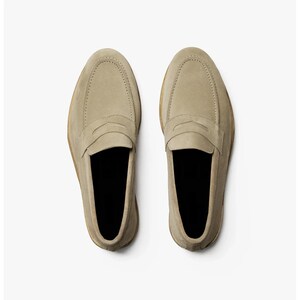 May include: A top-down view of two beige suede loafers. The shoes have a classic penny loafer design with a stitched vamp and a rounded toe. The soles are a light brown color, and the interior is black.