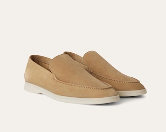 Puede incluir: Un par de mocasines de ante color canela con punta redondeada y suela blanca. Los zapatos tienen un diseño sin cordones y detalles de costura visibles. El calzado se presenta sobre un fondo neutro.