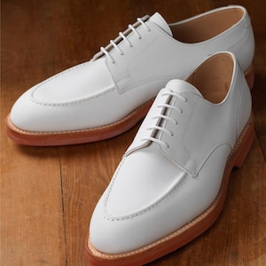 Puede incluir: Un par de zapatos de cuero blanco con un dise&ntilde;o cl&aacute;sico. Los zapatos tienen una punta redondeada, cierre con cordones y suela marr&oacute;n rojizo. Los zapatos se exhiben sobre una superficie de madera.