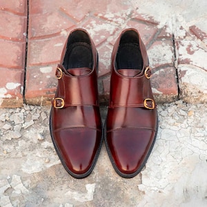 Peut inclure: Une paire de chaussures de ville en cuir bordeaux avec un design &agrave; double bride de moine. Les chaussures ont une finition polie, des bouts pointus et des boucles dor&eacute;es. Les chaussures sont pr&eacute;sent&eacute;es sur une surface en briques et en b&eacute;ton.