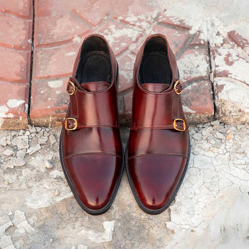 Peut inclure: Une paire de chaussures de ville en cuir bordeaux avec un design &agrave; double bride de moine. Les chaussures ont une finition polie, des bouts pointus et des boucles dor&eacute;es. Les chaussures sont pr&eacute;sent&eacute;es sur une surface en briques et en b&eacute;ton.