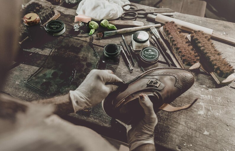 Pode incluir: Um artes&atilde;o trabalha num sapato de couro castanho com detalhes brogue. Ferramentas de cuidado de sapatos, incluindo escovas e cremes, est&atilde;o dispostas numa superf&iacute;cie de madeira, sugerindo uma repara&ccedil;&atilde;o ou restaura&ccedil;&atilde;o de sapatos.