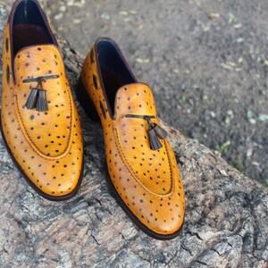 May include: A pair of yellow leather loafers with black dot detailing. Each shoe features a tassel accent and a dark brown sole. The shoes are displayed on a textured, natural surface.