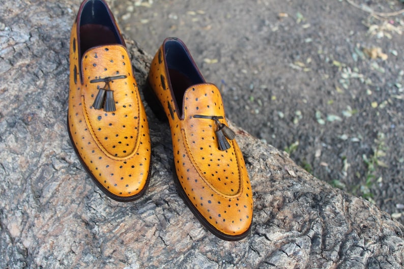 May include: A pair of yellow leather loafers with black dot detailing. Each shoe features a tassel accent and a dark brown sole. The shoes are displayed on a textured, natural surface.