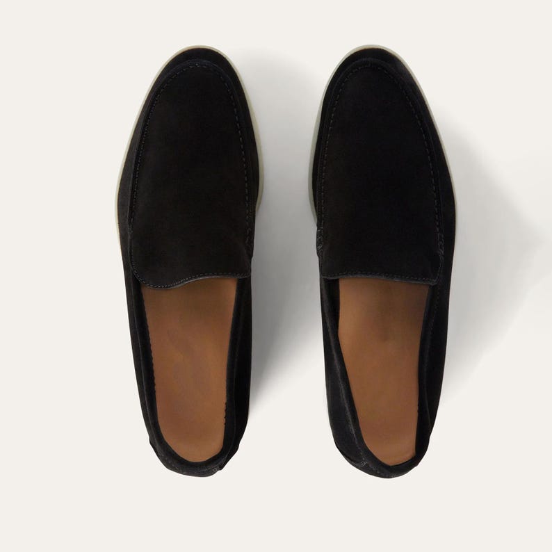 K&ouml;nnte beinhalten: Ein Paar schwarze Wildleder-Loafer mit runder Spitze und Slip-on-Design. Die Schuhe haben eine hellfarbene Sohle und ein braunes Innenfutter. Das Bild ist aus der Vogelperspektive aufgenommen und zeigt die Schuhe.