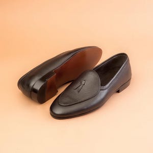 May include: Two black leather loafers with tassel detailing. The shoes have a rounded toe and a low heel. The soles are brown. The shoes are displayed on a light orange background. The shoes are made of leather.