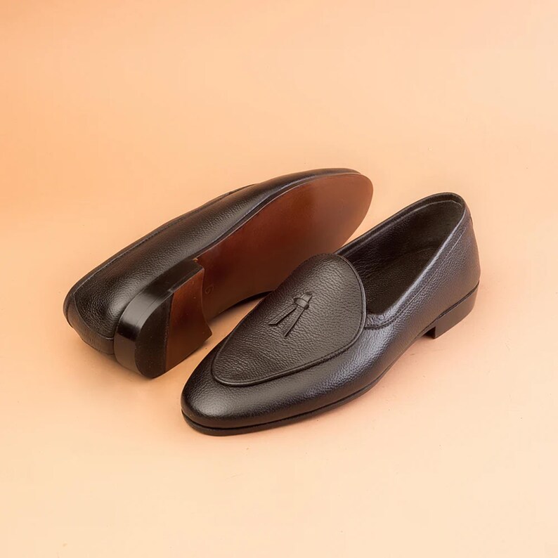 May include: Two black leather loafers with tassel detailing. The shoes have a rounded toe and a low heel. The soles are brown. The shoes are displayed on a light orange background. The shoes are made of leather.
