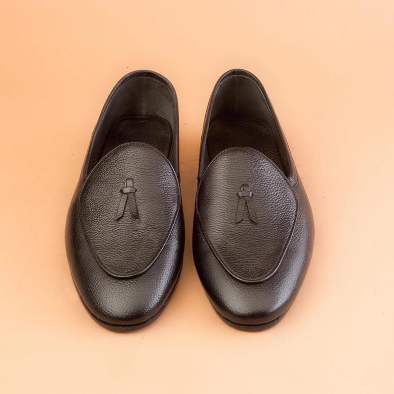 May include: A pair of black leather loafers with a textured finish. Each shoe features a decorative tassel detail on the vamp. The shoes are displayed on a light orange background.