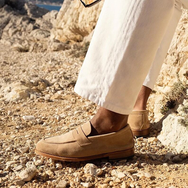 May include: Tan suede penny loafers with a leather sole. The shoes are worn with white pants. The image is taken outdoors on a rocky surface.