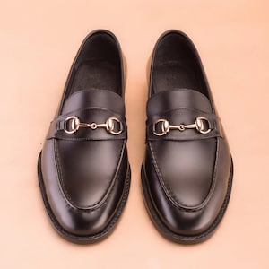K&ouml;nnte beinhalten: Ein Paar schwarze Leder-Loafer mit goldfarbenem Metall-Pferdegebiss-Detail. Die Schuhe haben ein klassisches Design mit runder Spitze und niedrigem Absatz. Die Loafer werden auf einer hellpfirsichfarbenen Oberfl&auml;che pr&auml;sentiert.