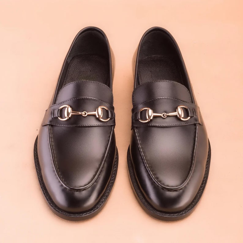 K&ouml;nnte beinhalten: Ein Paar schwarze Leder-Loafer mit goldfarbenem Metall-Pferdegebiss-Detail. Die Schuhe haben ein klassisches Design mit runder Spitze und niedrigem Absatz. Die Loafer werden auf einer hellpfirsichfarbenen Oberfl&auml;che pr&auml;sentiert.