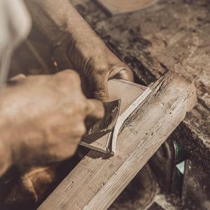 K&ouml;nnte beinhalten: Ein Handwerker bearbeitet mit einem Handhobel ein St&uuml;ck Holz. Das Holz ist hellbraun, und die H&auml;nde des Handwerkers sind bei der Arbeit zu sehen. Die Szene spielt in einer Werkstatt mit strukturierter Oberfl&auml;che.