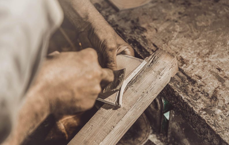 K&ouml;nnte beinhalten: Ein Handwerker bearbeitet mit einem Handhobel ein St&uuml;ck Holz. Das Holz ist hellbraun, und die H&auml;nde des Handwerkers sind bei der Arbeit zu sehen. Die Szene spielt in einer Werkstatt mit strukturierter Oberfl&auml;che.