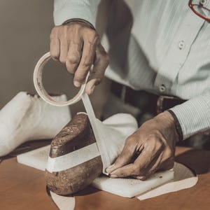 Pode incluir: Um sapateiro aplica fita adesiva a uma forma de sapato. A cena mostra uma forma de sapato de couro castanho, uma forma de sapato branca e um rolo de fita adesiva. O sapateiro est&aacute; a usar uma camisa azul clara &agrave;s riscas.