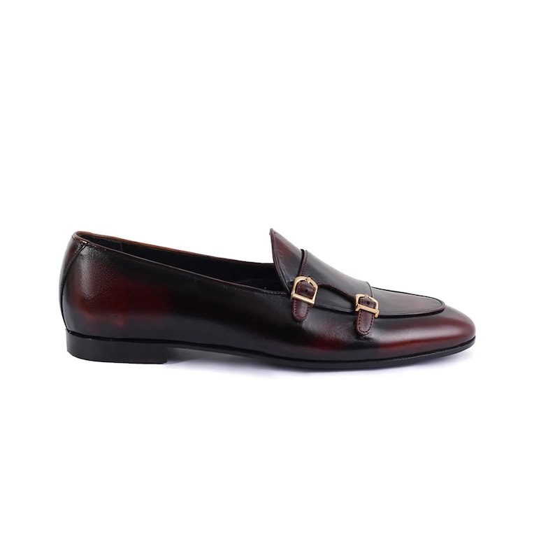 May include: A single, burgundy leather loafer with a double monk strap closure. The shoe features a sleek design with a polished finish and gold-colored buckles. The shoe is designed for formal or business attire.