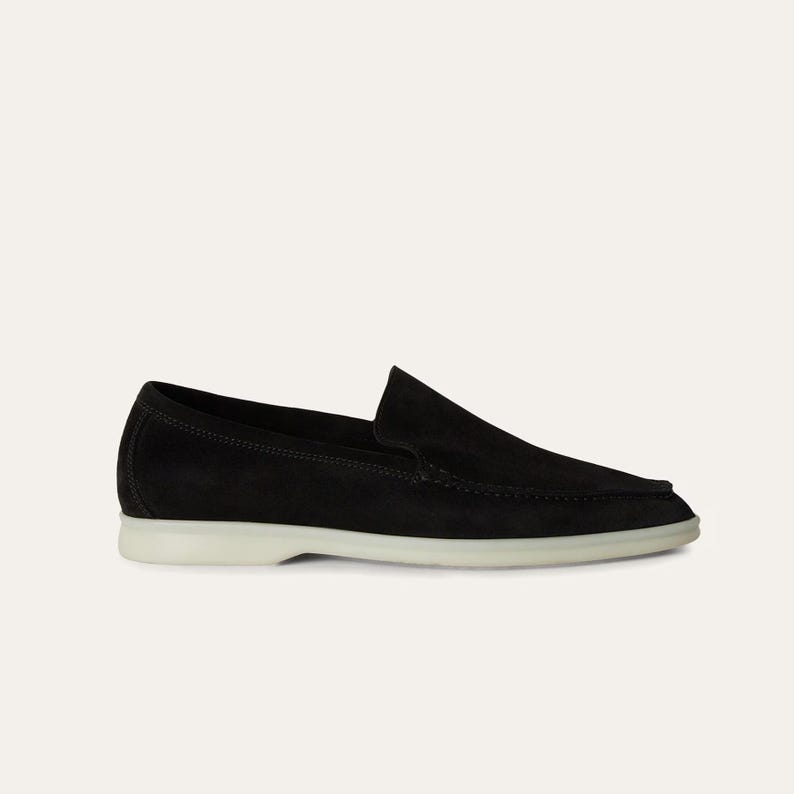 K&ouml;nnte beinhalten: Schwarzer Wildleder-Loafer mit wei&szlig;er Sohle. Der Schuh hat ein klassisches Slip-on-Design mit runder Spitze und niedrigem Absatz. Dieses Schuhwerk eignet sich f&uuml;r l&auml;ssige oder halbformelle Anl&auml;sse.