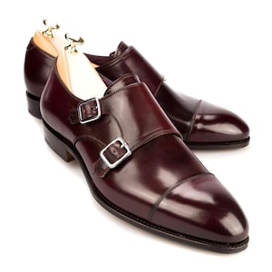 Pode incluir: Um par de sapatos de couro bordô com fivelas duplas e fivelas prateadas. Os sapatos têm um acabamento polido e são exibidos com formas de madeira no interior. As biqueiras são de um tom bordô mais escuro.