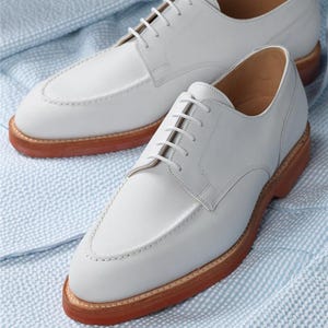 Puede incluir: Un par de zapatos de vestir de cuero blanco con un dise&ntilde;o cl&aacute;sico. Los zapatos tienen cierre de cordones, puntera cosida y suela marr&oacute;n. Se exhiben sobre una tela estampada en azul claro y blanco.