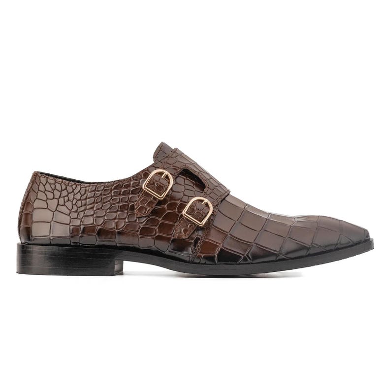 K&ouml;nnte beinhalten: Brauner Lederschuh mit Krokodiltextur und spitzer Zehenpartie. Der Schuh hat zwei goldfarbene Schnallen und eine schwarze Sohle. Ein klassischer Stil f&uuml;r formelle Anl&auml;sse.