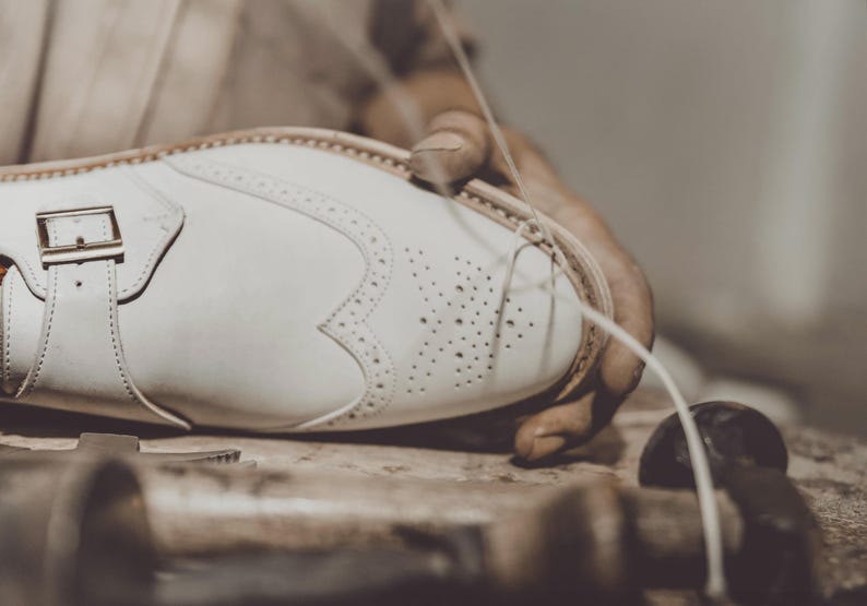 K&ouml;nnte beinhalten: Nahaufnahme eines wei&szlig;en Lederschuhs, der von Hand gen&auml;ht wird. Der Schuh hat dekorative Perforationen, eine Schnalle und einen Riemen. Das Bild zeigt die Handwerkskunst der Schuhmacherei.