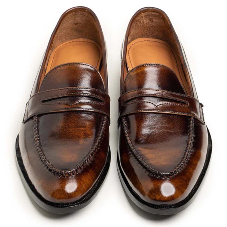 May include: A pair of brown leather penny loafers. The shoes have a glossy finish, a stitched vamp, and a strap across the top. The soles are black. The shoes are displayed on a white background.