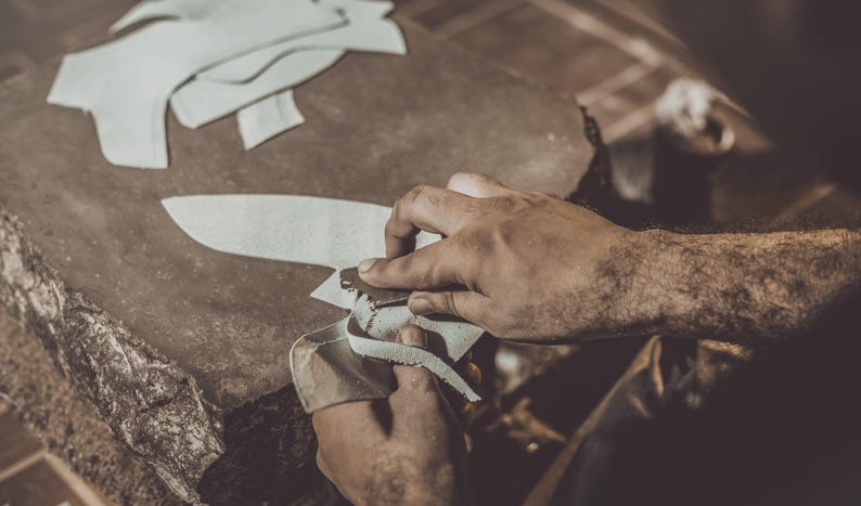 K&ouml;nnte beinhalten: Nahaufnahme einer Person, die mit Leder arbeitet. Die Person verwendet eine Klinge, um ein St&uuml;ck wei&szlig;es Leder zu schneiden. Mehrere zugeschnittene Lederst&uuml;cke liegen auf der Arbeitsfl&auml;che. Warme Farbt&ouml;ne.