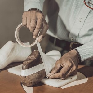 May include: A person is shown working on a shoe form, applying tape. The scene is set in a workshop, with a white shoe form and a brown leather shoe form visible. A knife and tape roll are on the table.
