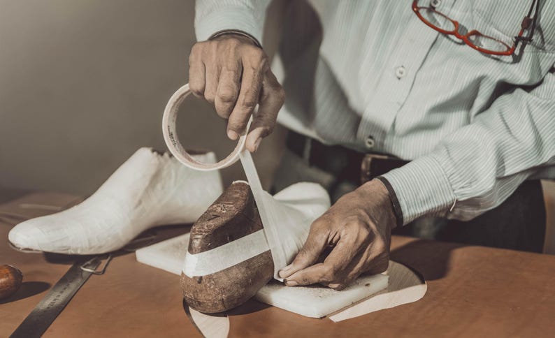 May include: A person is shown working on a shoe form, applying tape. The scene is set in a workshop, with a white shoe form and a brown leather shoe form visible. A knife and tape roll are on the table.