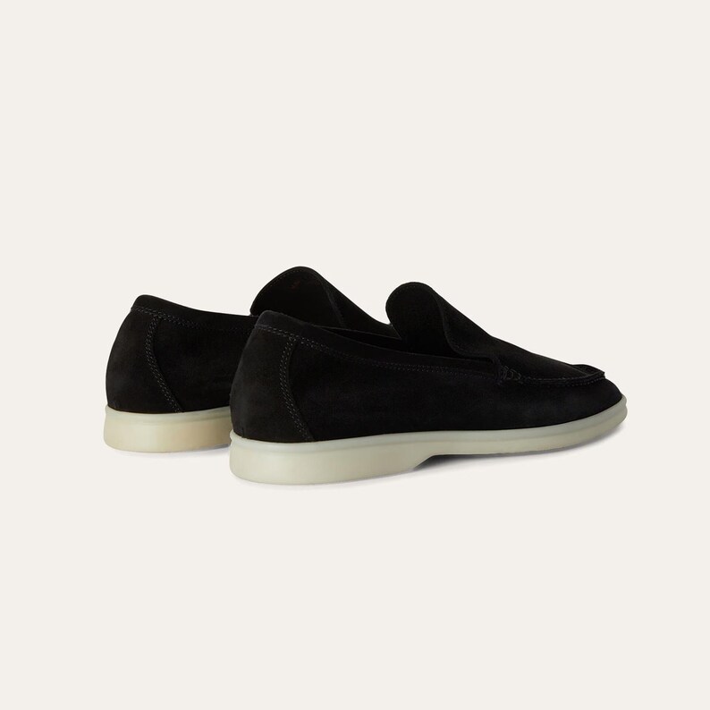 K&ouml;nnte beinhalten: Ein Paar schwarze Wildleder-Loafer mit Slip-on-Design. Die Schuhe haben eine hellfarbige Sohle und eine runde Spitze. Das Bild zeigt die Schuhe von der Seite, wobei die Textur und Form der Schuhe hervorgehoben werden.