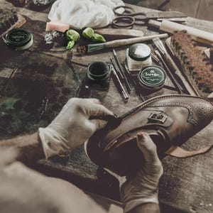 May include: A person wearing gloves is working on a brown leather shoe with brogue detailing. The shoe is on a wooden surface with various shoe care tools, including brushes, creams, and applicators. The scene suggests shoe repair or restoration.
