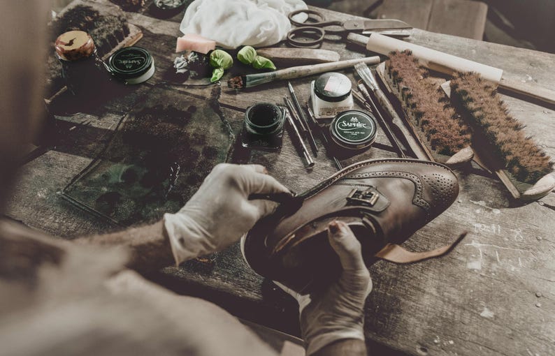 May include: A person wearing gloves is working on a brown leather shoe with brogue detailing. The shoe is on a wooden surface with various shoe care tools, including brushes, creams, and applicators. The scene suggests shoe repair or restoration.