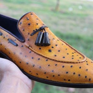 May include: A tan leather loafer with a distinctive dotted pattern and a black tassel detail. The shoe has a dark brown sole and a navy blue interior. The shoe is held by a hand against a blurred green background.