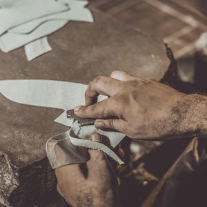 May include: A close-up shot of a person working with leather. The person is using a blade to cut a piece of white leather. Several cut leather pieces are on the work surface. The scene is lit with warm tones.