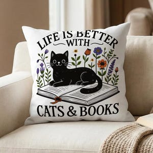 Könnte beinhalten: Weißes quadratisches Kissen mit dem Text "LIFE IS BETTER WITH CATS & BOOKS". Es zeigt eine schwarze Katze, die auf einem offenen Buch liegt, umgeben von Blumen. Das Kissen liegt auf einem beigen Sofa.