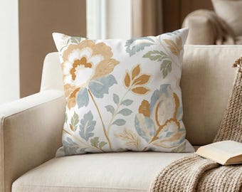 May include: Square decorative pillow with a floral pattern in shades of gold, blue, and green on a white background. The pillow is placed on a beige sofa, with a knitted throw and an open book nearby.