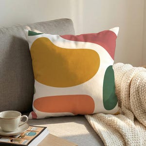 Puede incluir: Un cojín decorativo con un diseño abstracto. El cojín presenta grandes formas irregulares en amarillo mostaza, coral, verde y rojo sobre un fondo blanco. El cojín está sobre un sofá gris con una manta color crema.