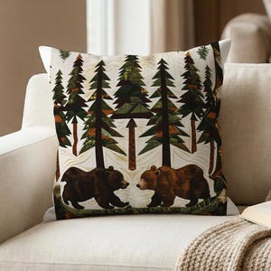 May include: Square decorative pillow with a forest scene featuring two brown bears facing each other. The pillow has a white background with green and brown trees and a quilted design. The pillow is on a beige sofa.