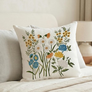 May include: White square pillow with an embroidered floral design. The embroidery features a variety of colorful flowers, including daisies, blue hydrangeas, and yellow blooms. The pillow is placed on a bed with other pillows.