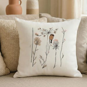 May include: Cream-colored square throw pillow featuring a delicate illustration of a bird perched on a floral stem. The design includes black line art and soft pink and brown accents, creating a nature-inspired aesthetic. The pillow is placed on a beige sofa.