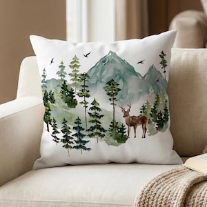 May include: White square throw pillow with a watercolor forest scene. The design features green trees, a mountain range, a deer, and flying birds. The pillow is placed on a beige sofa.