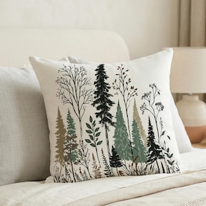 Pode incluir: Uma almofada quadrada branca com um desenho de floresta. O design apresenta árvores em tons de verde, castanho e preto, com ramos e folhas detalhadas. A almofada está colocada numa cama, juntamente com outras almofadas.