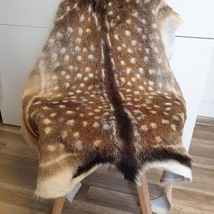 Pode incluir: Uma pele de veado com manchas castanhas e brancas, drapeada sobre um banco de madeira. A pele tem um padr&atilde;o natural com detalhes em castanho escuro e preto. O banco tem quatro pernas de madeira.
