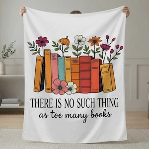 May include: White blanket with a colorful illustration of books and flowers. The design includes a row of books in various colors with flowers sprouting from the tops. The text reads, "THERE IS NO SUCH THING as toe many books."