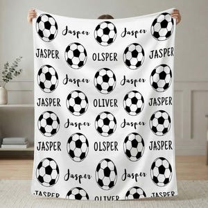 May include: White blanket featuring a repeating pattern of black and white soccer balls and names. The names "Jasper" and "Oliver" are written in a cursive font. The blanket is held up by a person.