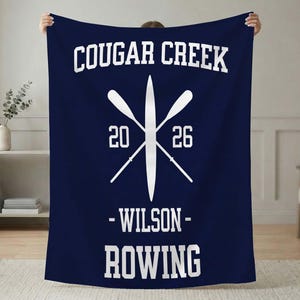 May include: A navy blue blanket with white text and a rowing theme. The blanket features the words "COUGAR CREEK" above a crossed oar design with the numbers "20" and "26". Below the design are the words "- WILSON - ROWING".