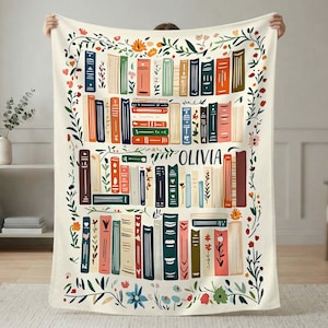 Puede incluir: Una manta crema con un diseño de estantería con libros coloridos y detalles florales. El nombre "OLIVIA" está impreso en el centro. La manta está sostenida por una persona.