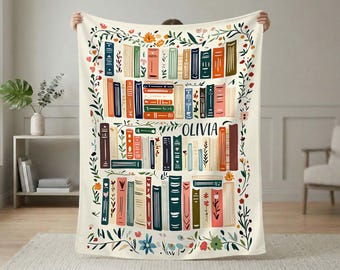 Könnte beinhalten: Eine cremefarbene Wohndecke mit einer bunten Illustration von Bücherstapeln in Regalen, durchsetzt mit floralen Ranken und Blättern. Der Name OLIVIA ist in dunkelblauen Großbuchstaben zwischen den Buchrücken aufgedruckt.