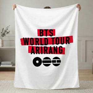 Puede incluir: Manta blanca con el texto "BTS WORLD TOUR ARIRANG" en negro, sobre bloques rectangulares rojos. Debajo del texto hay tres diseños circulares negros. La manta está sostenida por una persona.